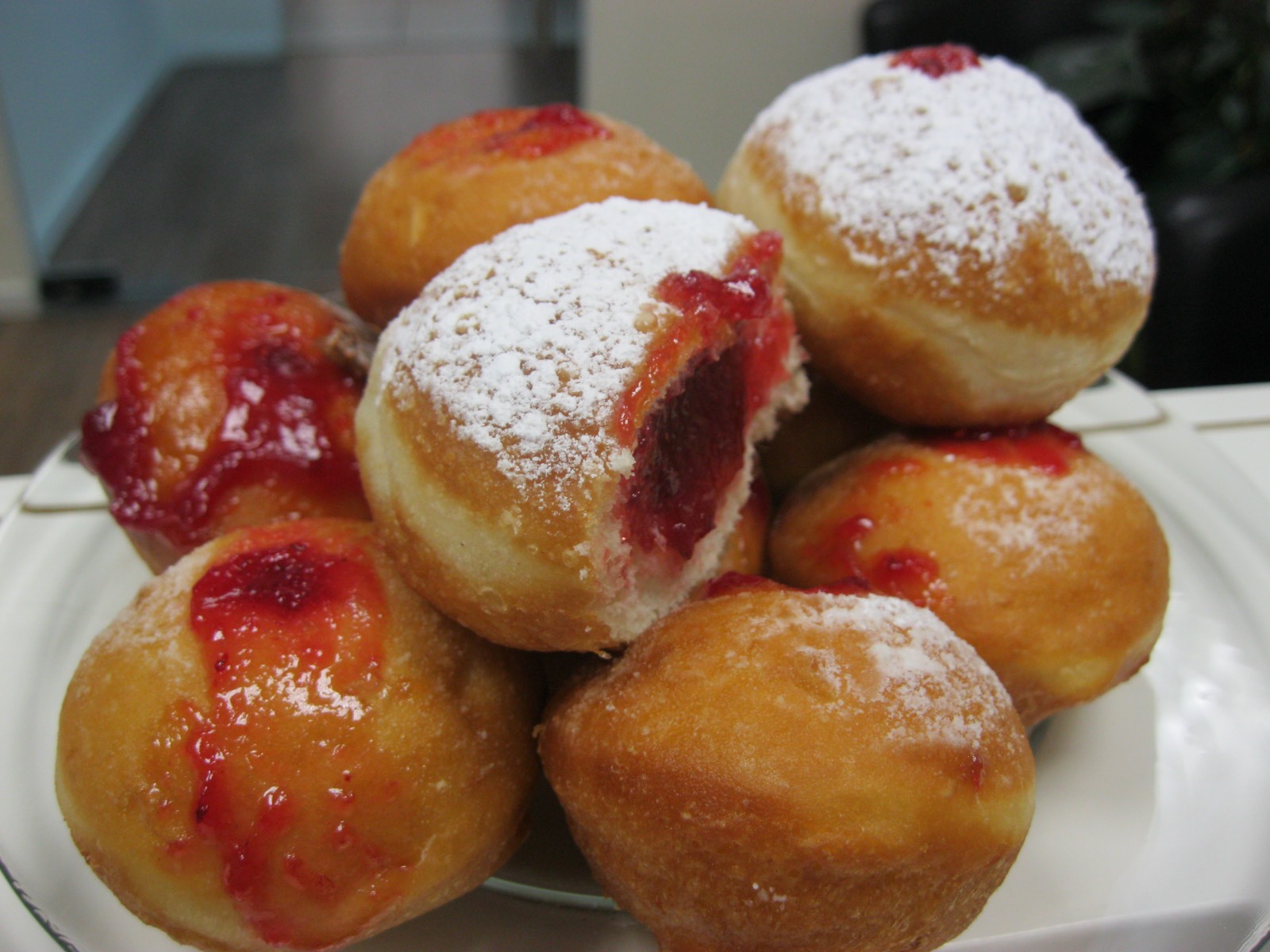 A plate of sufganiyot, golden jelly doughnuts dusted with powdered sugar, one bitten to show the red jam inside
